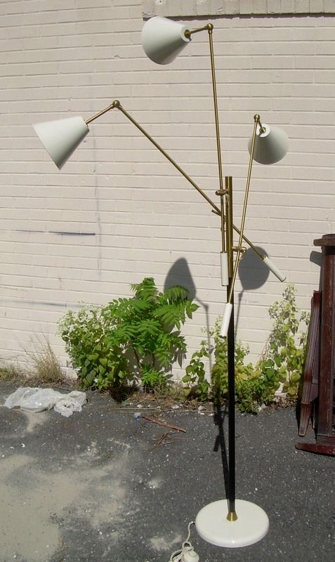 Tall brass floor lamp with three white shades on a white base outdoors against a brick wall.