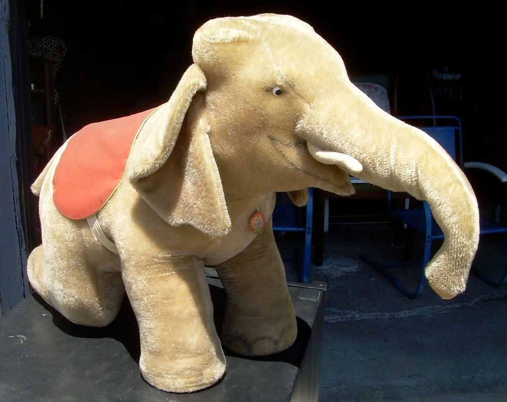 Plush elephant with a red saddle standing on a dark floor indoors
