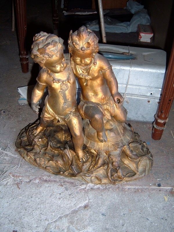 Gold cherub figurines on a textured base, displayed on a floor beside furniture and a silver case