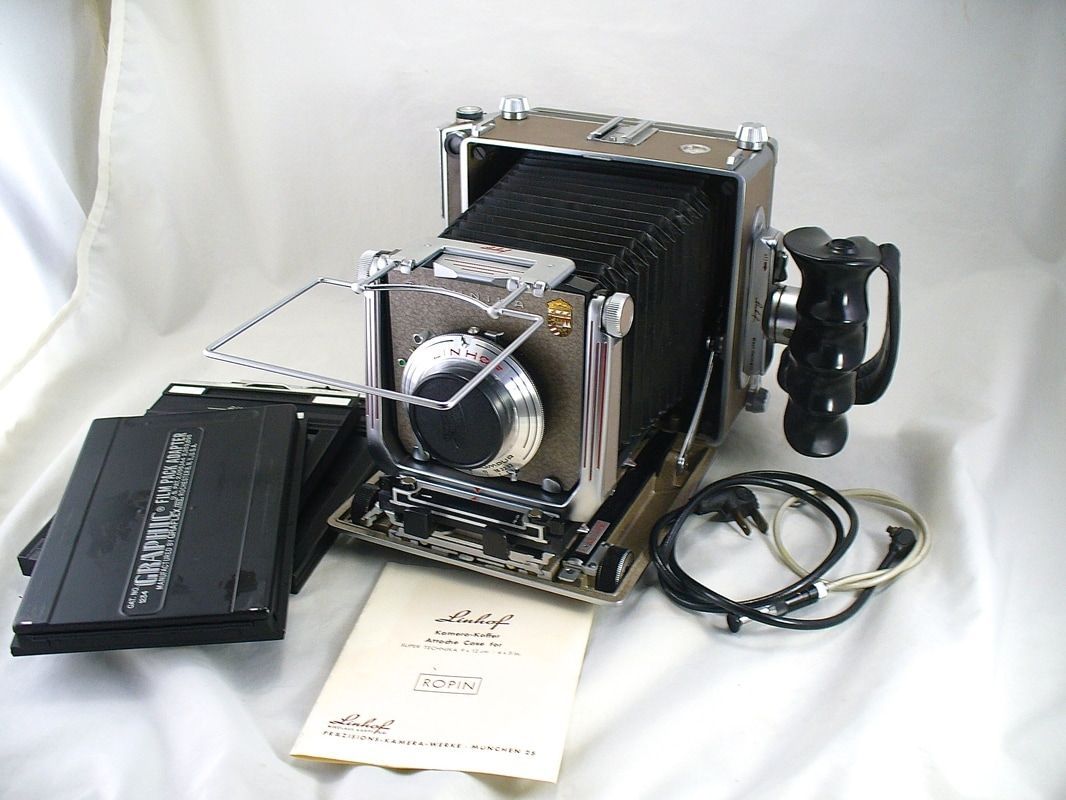 Vintage large-format camera kit with bellows, lens, carrying case, cables, and instruction leaflet on white background