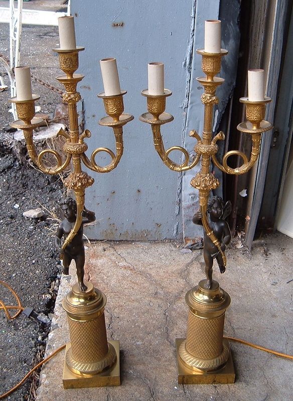 Pair of ornate gold candelabra lamps with white shades, standing on a rough floor near a blue door.