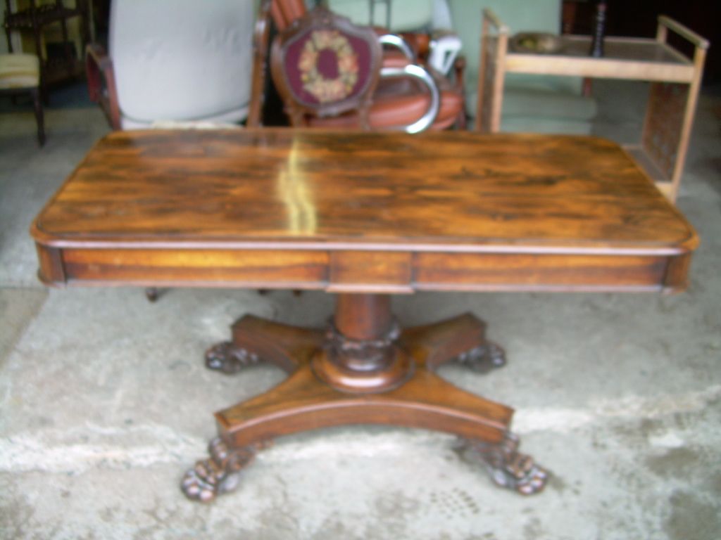 Wooden dining table with a polished surface and carved pedestal base on a concrete floor.