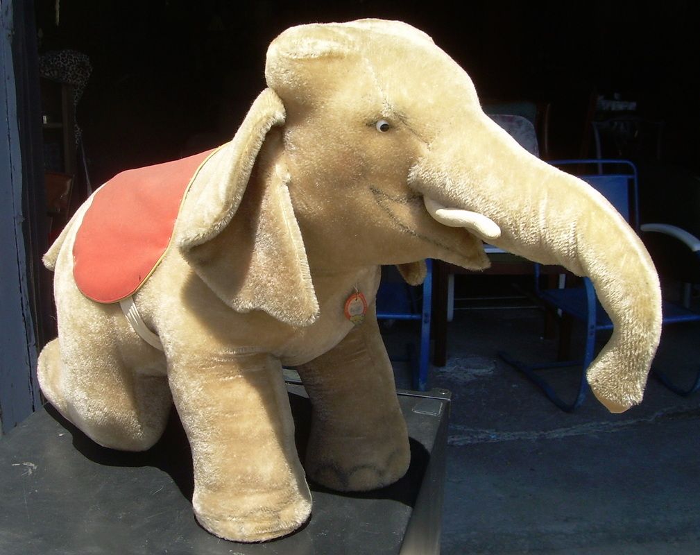 Beige plush elephant with red saddle standing indoors on a dark floor