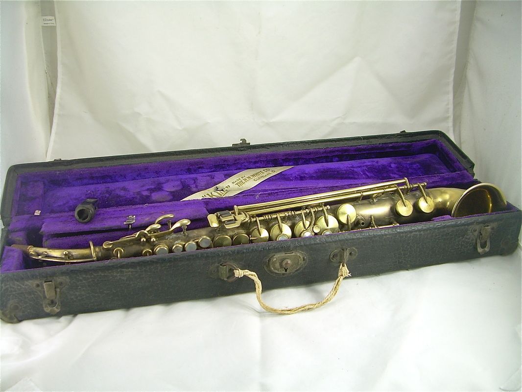 Open black instrument case lined with purple velvet, holding a gold saxophone mouthpiece and accessories.