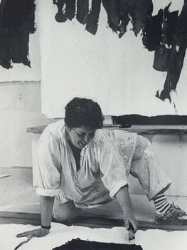 Person kneeling on the floor, drawing on paper in front of a large abstract black-and-white canvas.