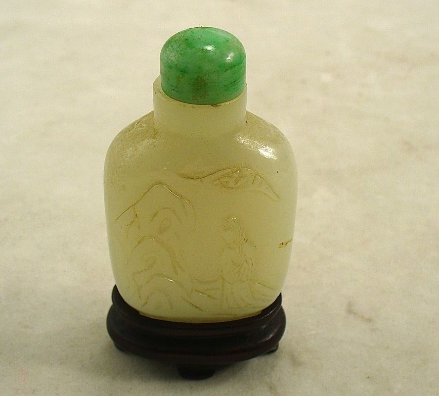 Small pale yellow carved bottle with a green cap, displayed on a dark wooden stand.