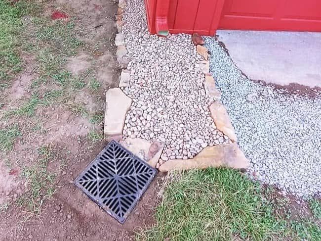 Drain beside red shed with gravel path, grass, and a black grate in the foreground
