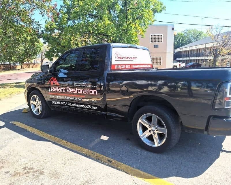Black pickup truck with “Reliant Restoration” signage parked beside a building in daylight