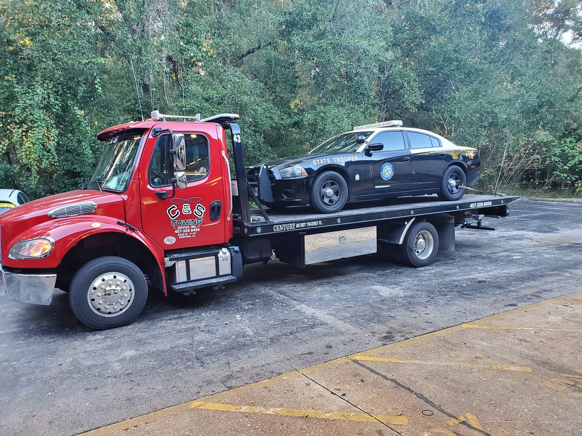 Red tow truck carrying a black police car on a flatbed trailer in a parking lot.