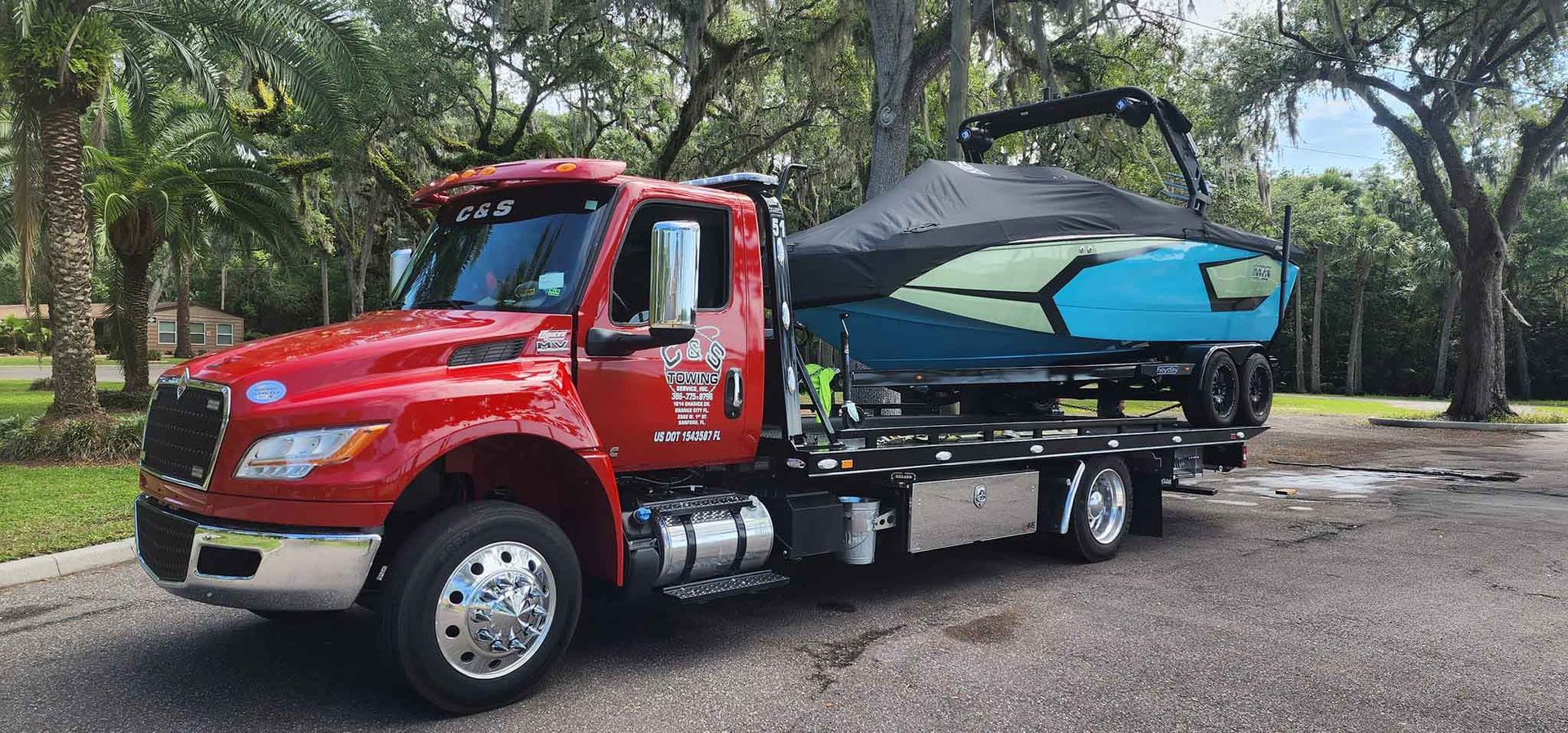 Red tow truck carrying a blue boat on a trailer in a park-like setting.