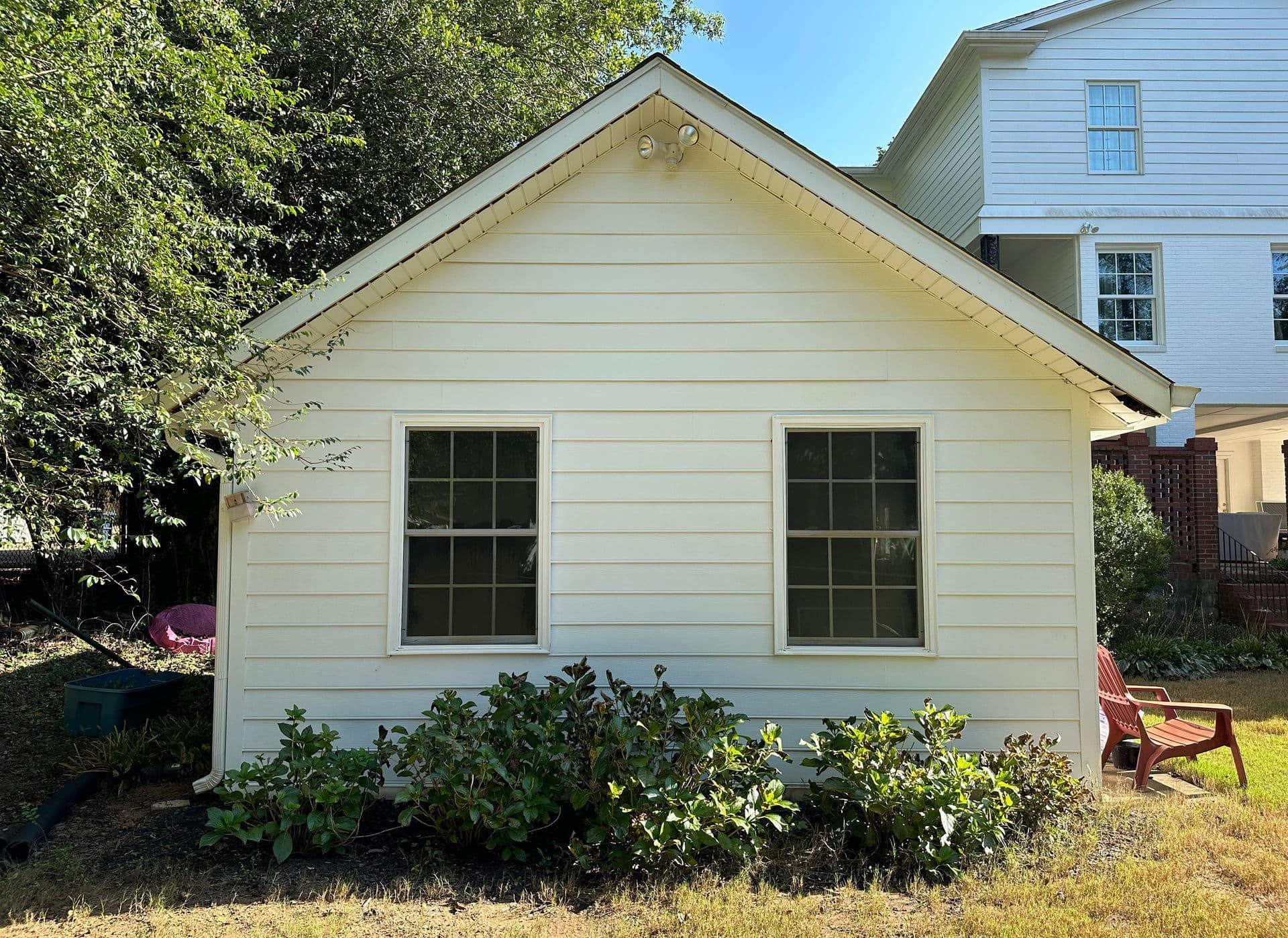 A small white house with two windows is surrounded by bushes and trees.