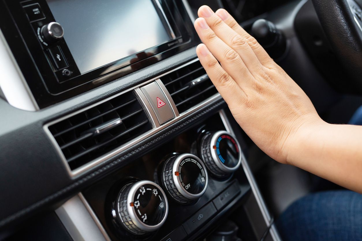 Hand hovering near a car dashboard air vent and climate controls with hazard button.