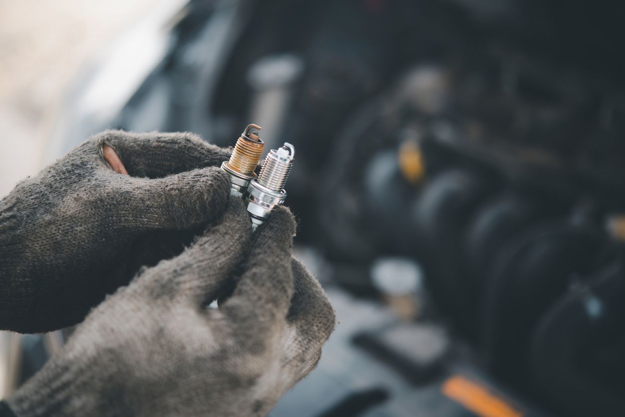 Gloved hands holding a small lit spark plug in a workshop, with a blurred engine in the background