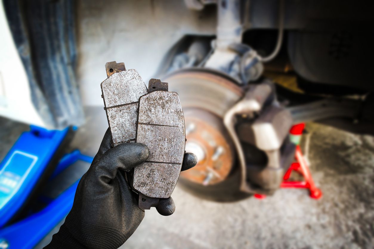 Gloved hand holding worn brake pads beside a car’s exposed brake rotor in a garage