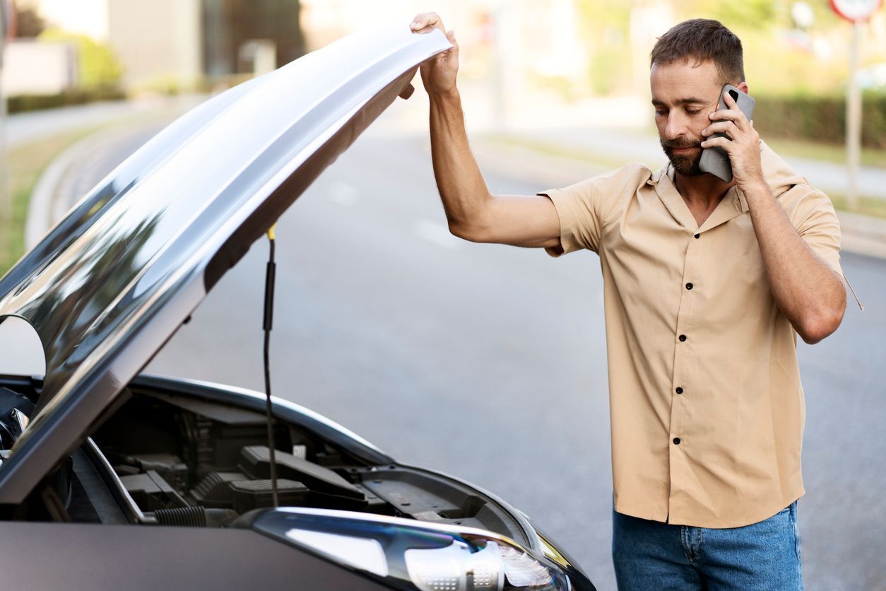 Man holding up a car hood while talking on a phone beside a broken-down vehicle