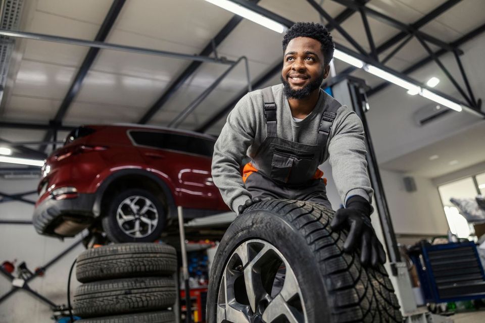 Mechanic smiling beside stacked tires in an auto shop, with a red SUV on a lift in the background
