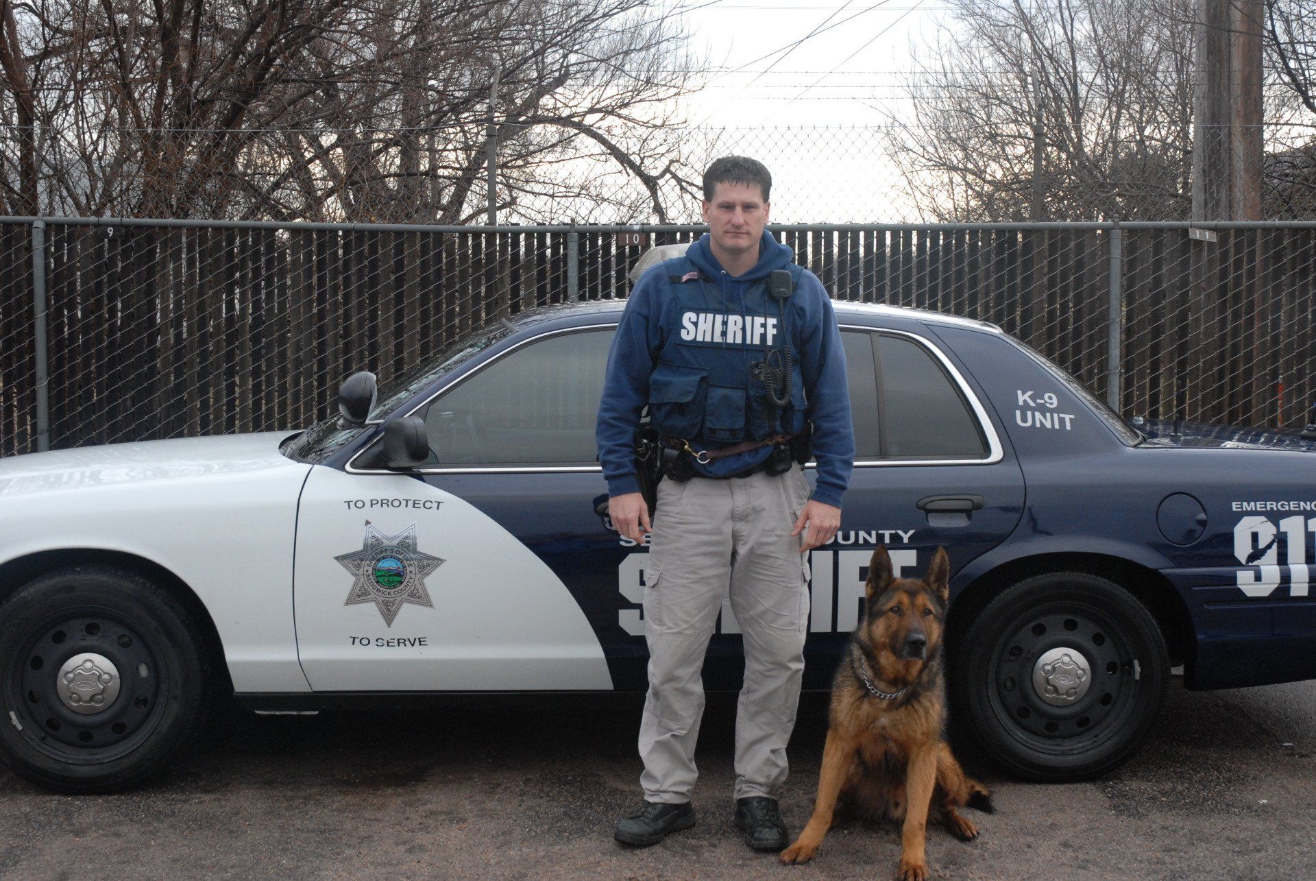 A sheriff stands next to a dog in front of a sheriff 's car