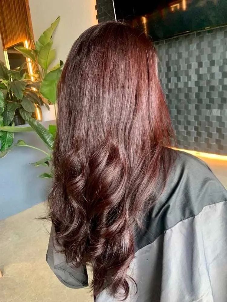 Long wavy auburn hair viewed from behind in a softly lit indoor setting with plants.