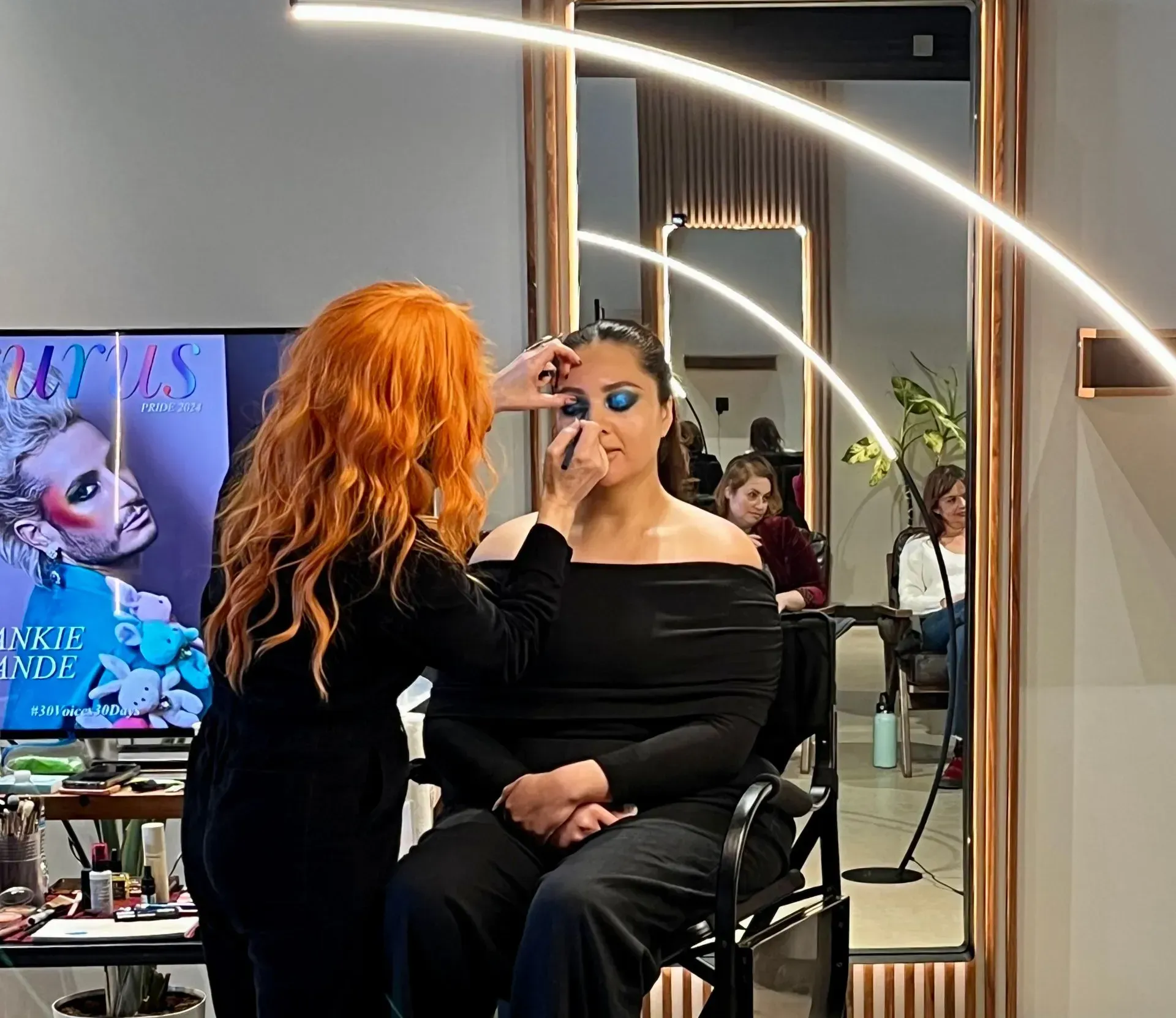 Woman getting makeup applied in a salon, seated before a mirror with a ring light.