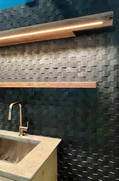 Modern kitchen with textured black tile wall, wood shelves, and a brass faucet over a stone sink.