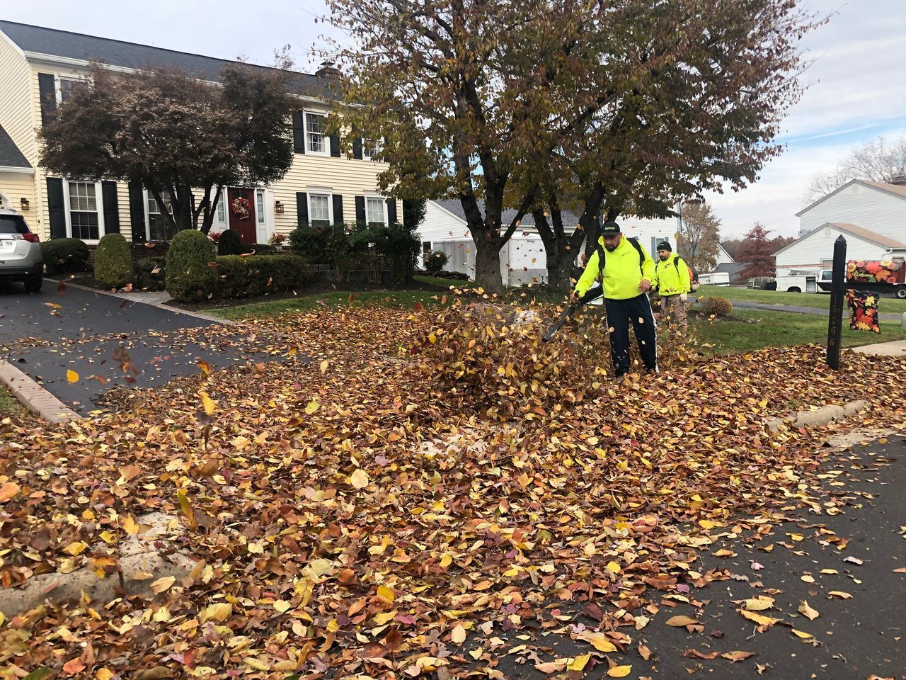 Workers clearing fallen leaves on a suburban street with bright yellow safety jackets