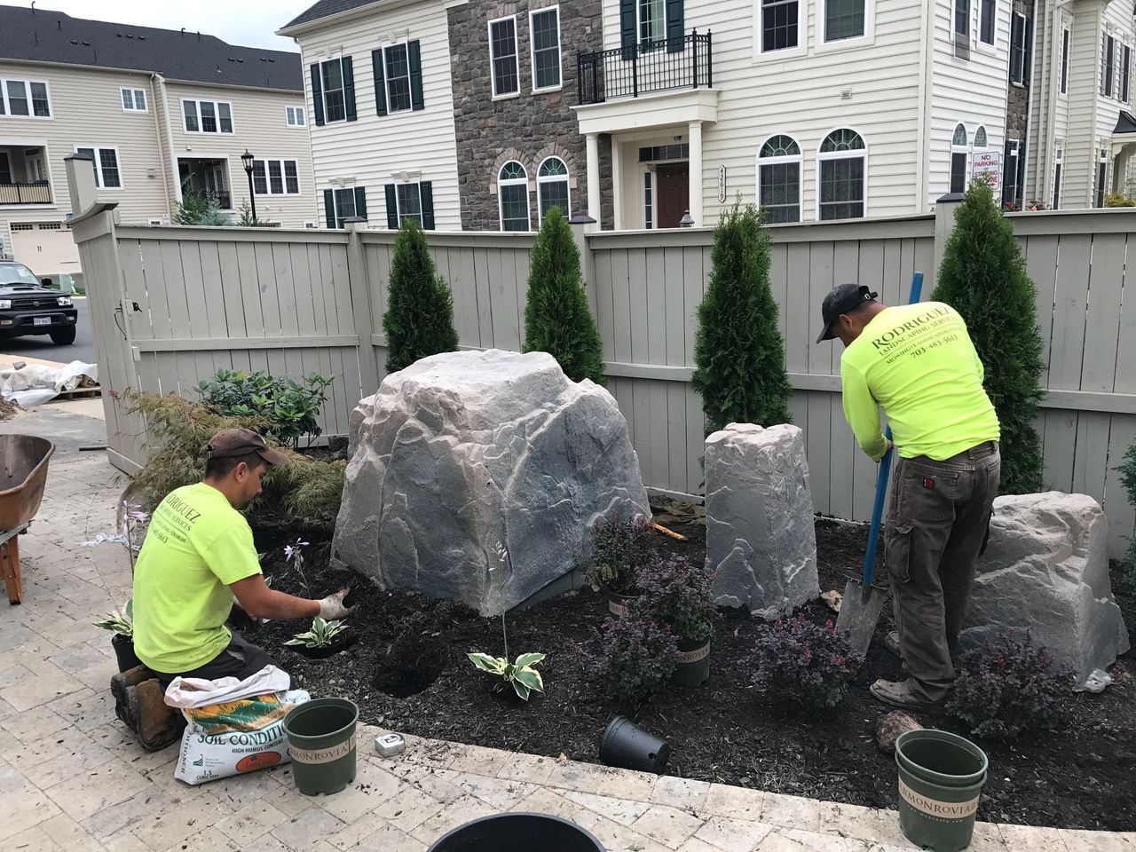 Two workers planting shrubs around large rocks in a landscaped front yard at a stone house.