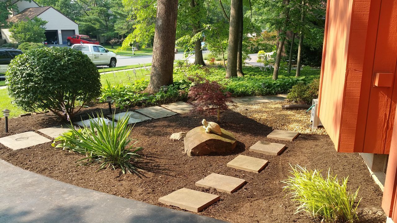 Landscaped garden bed beside a red house with stepping stones, mulch, shrubs, and trees in sunlight.