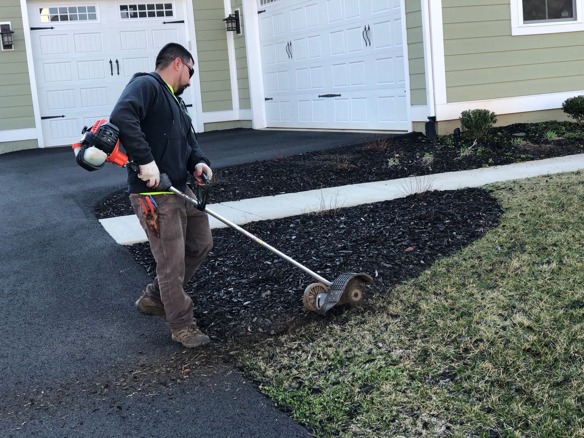 Person using a power broom to clear black driveway material beside a house and garage.