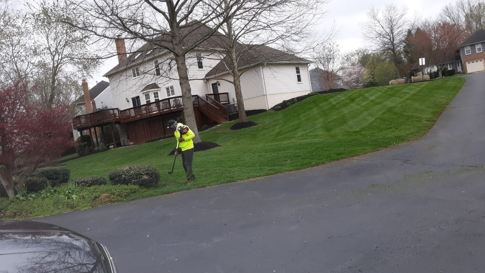 Person in a yellow jacket stands near a house on a sloped lawn beside a driveway.