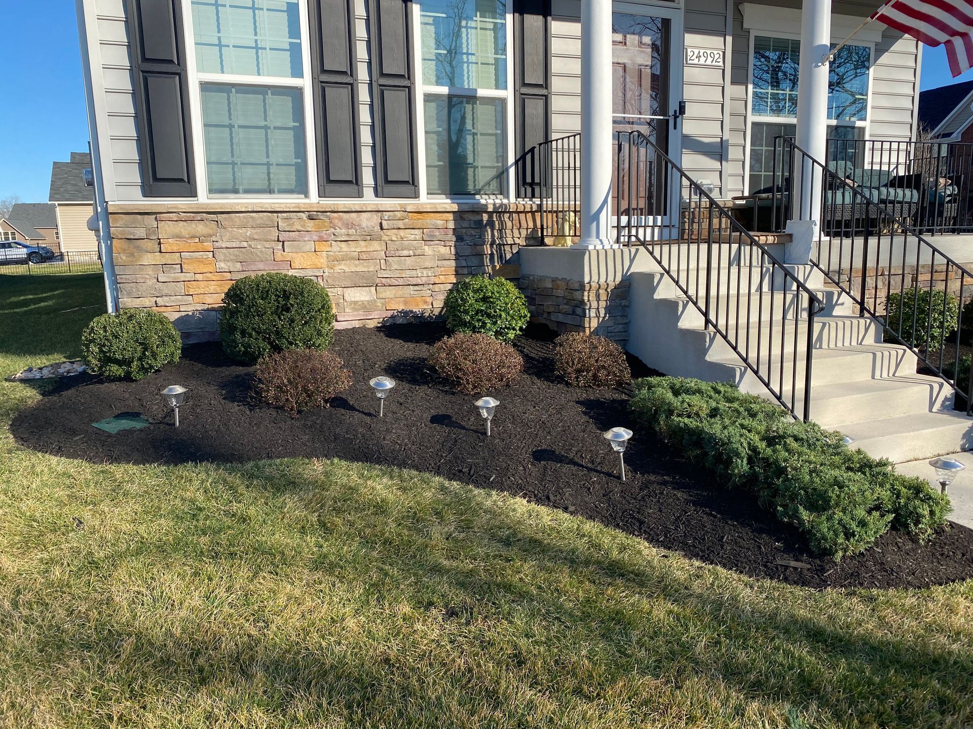 Front yard landscaping with dark mulch, shrubs, and stone steps beside a house with gray siding and stone facade