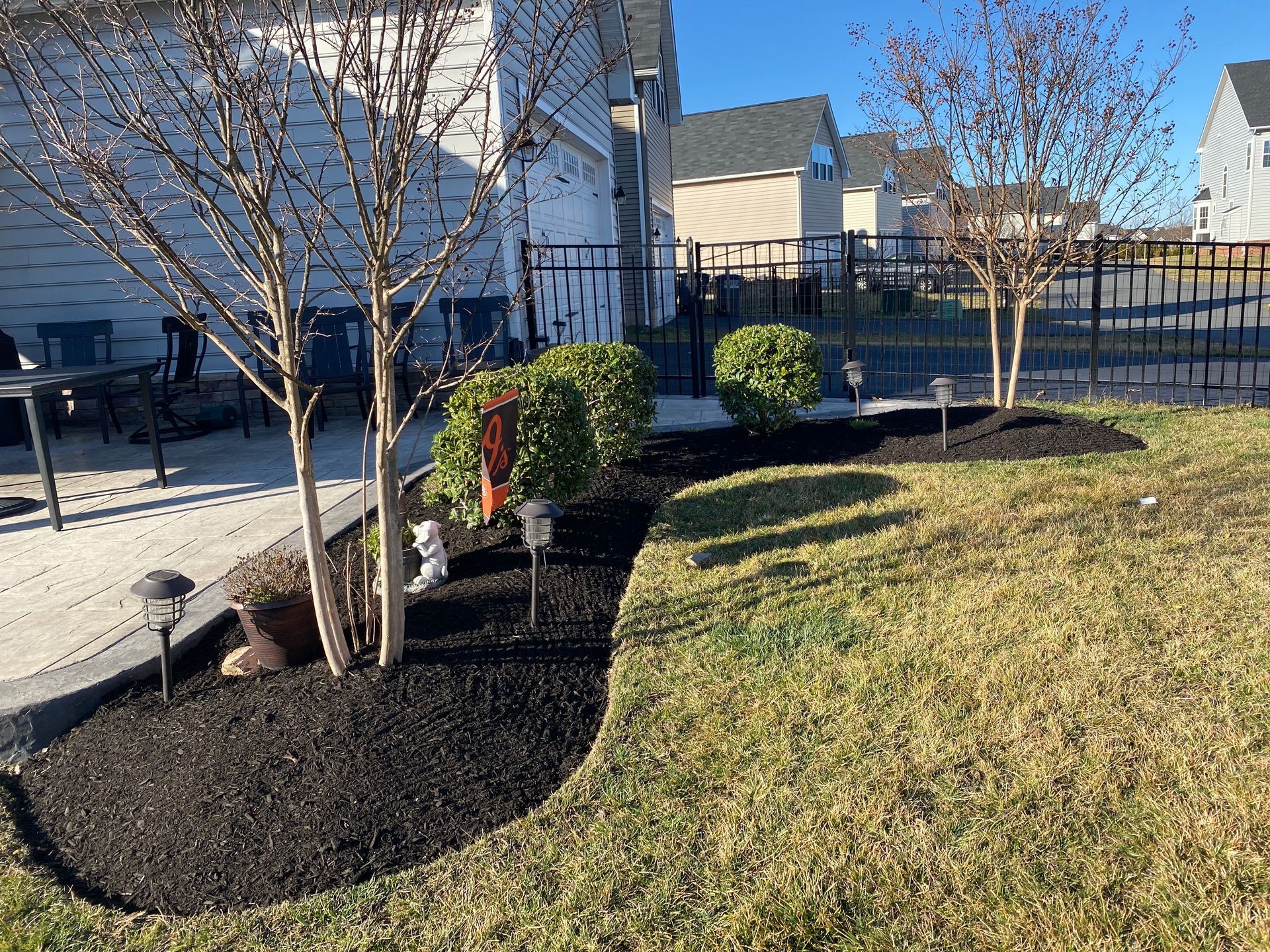 Residential front yard with young trees, mulch beds, shrubs, and a grassy lawn beside a house