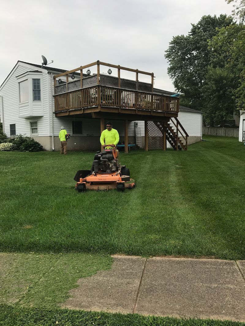 Two workers mowing a steep green lawn beside a white house with a wooden deck