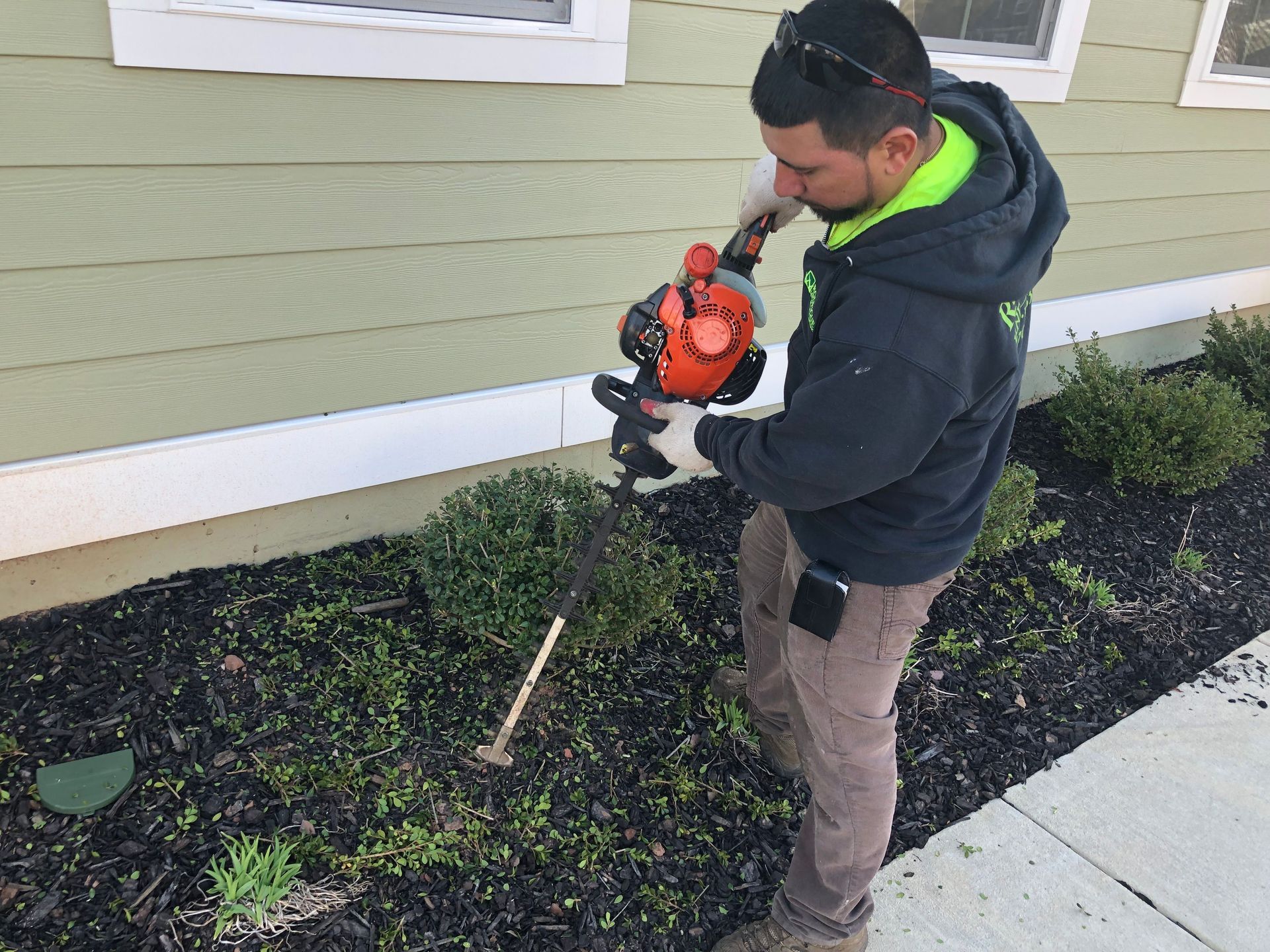 Person using a string trimmer beside a house, trimming weeds along a garden bed and sidewalk.