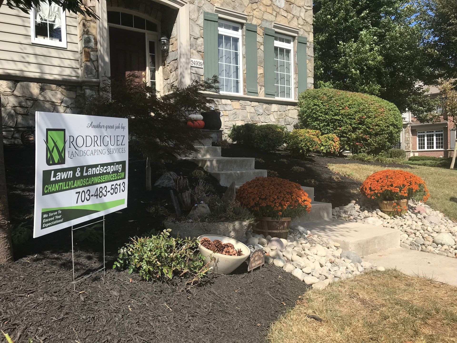 Landscape sign in front of a stone house with trimmed shrubs and orange flowers