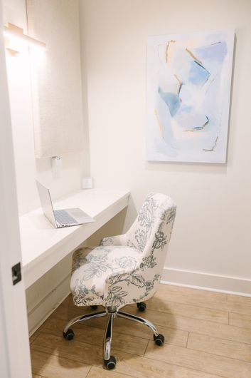 Desk with patterned chair, laptop, and abstract blue art on a beige wall, in a light-floored room.
