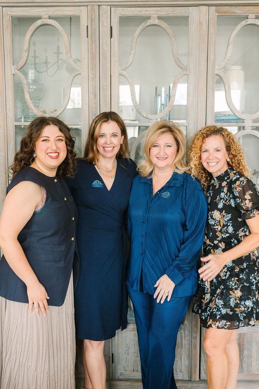 Four women smiling in front of a cabinet, all wearing blue or navy clothing.