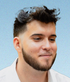 A young man with a beard is wearing a white shirt and smiling.