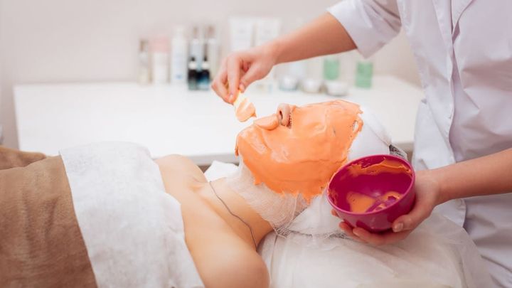 A person applying an orange facial mask while holding a bowl in a spa setting