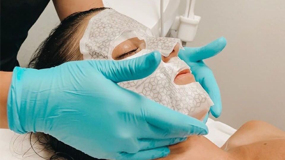 Gloved hands apply a white facial mask to a woman lying back during a spa treatment