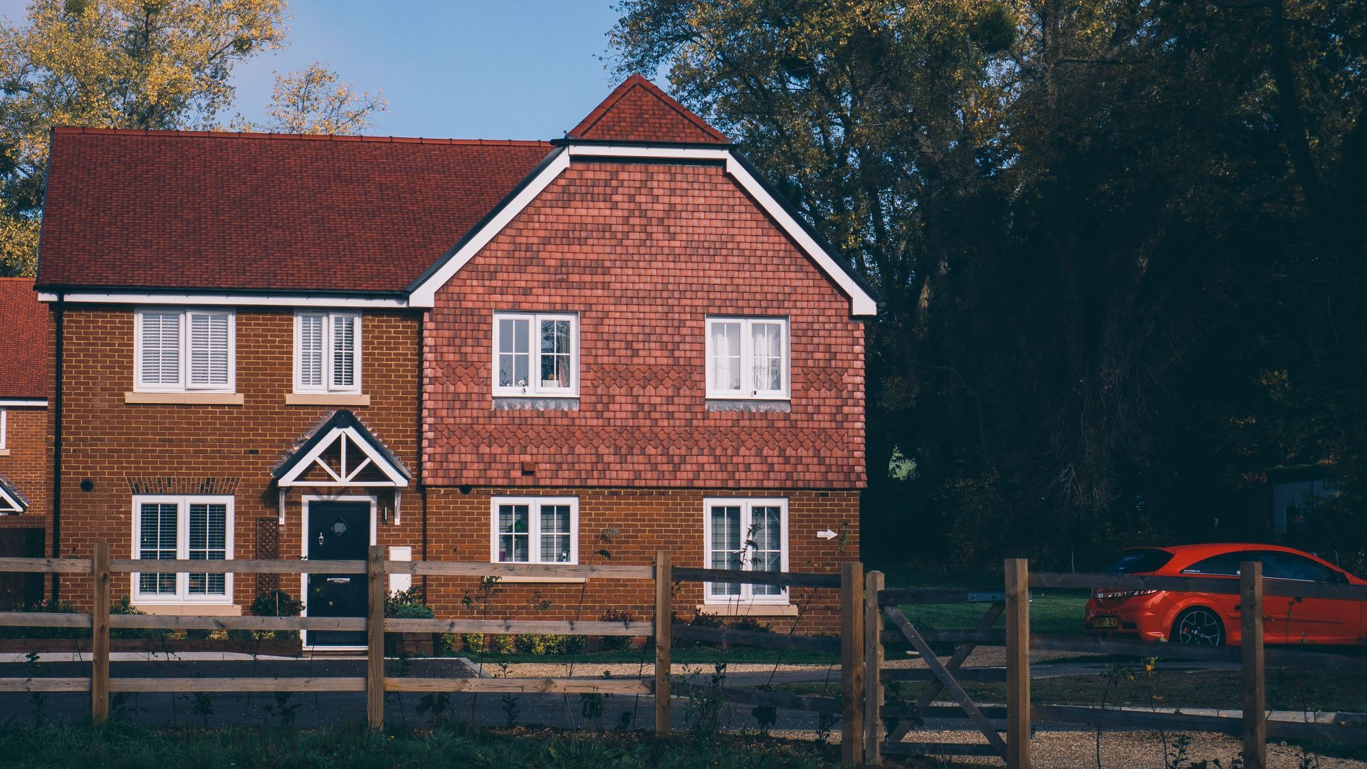 Red brick house with white windows and a front fence, with a red car parked on the right.