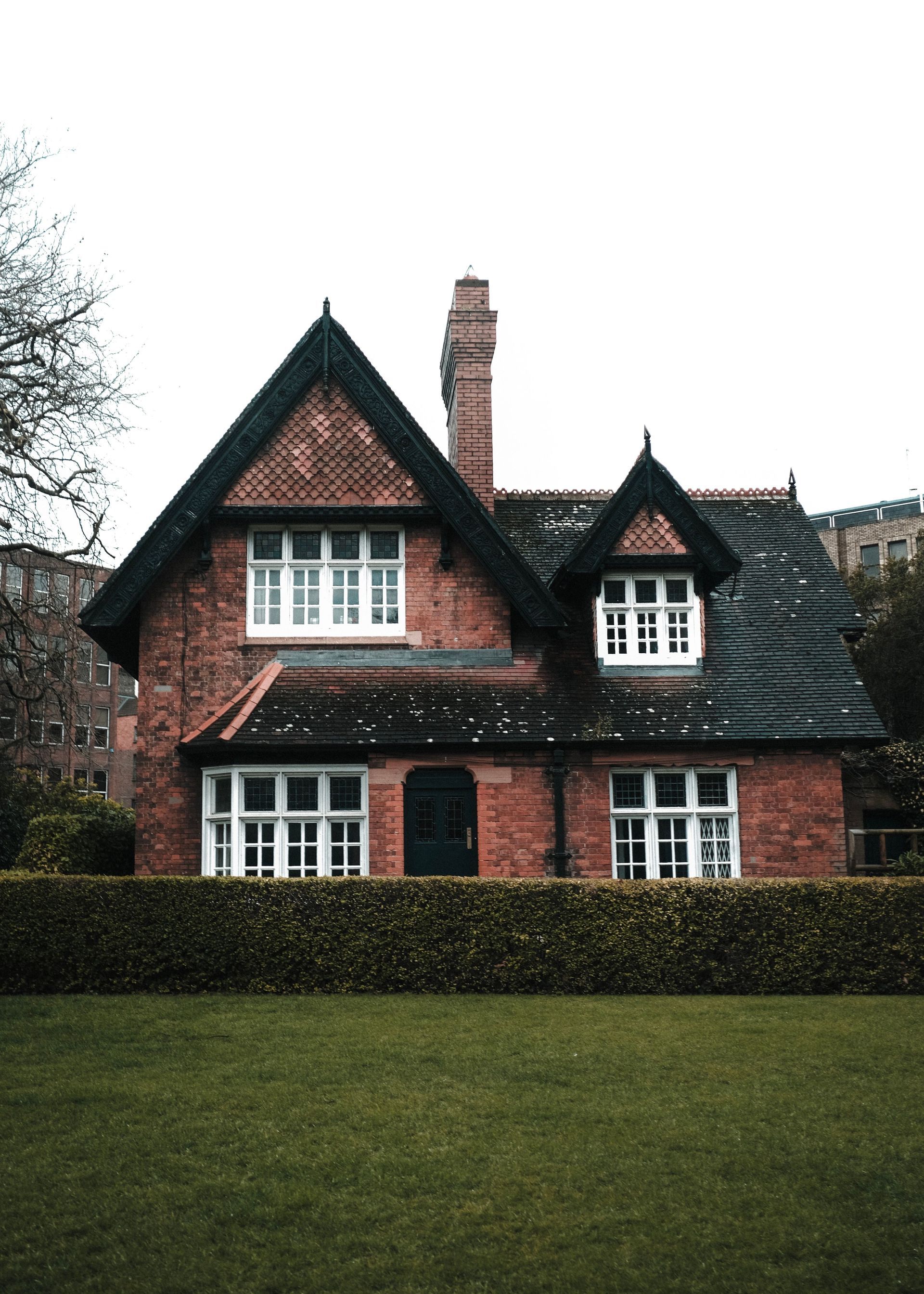Red brick house with dark roof, white-trimmed windows, and a hedge in front of a green lawn.