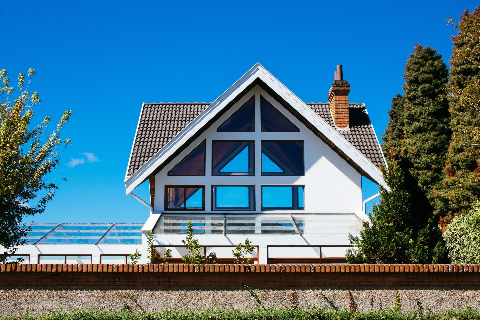 White house with blue windows and a steep roof under a clear blue sky, beside a brick wall and trees