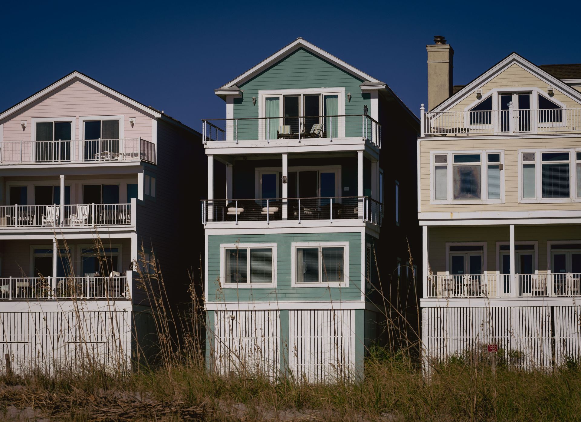 Three beach houses with porches, painted white and teal, behind dune grass under a blue sky