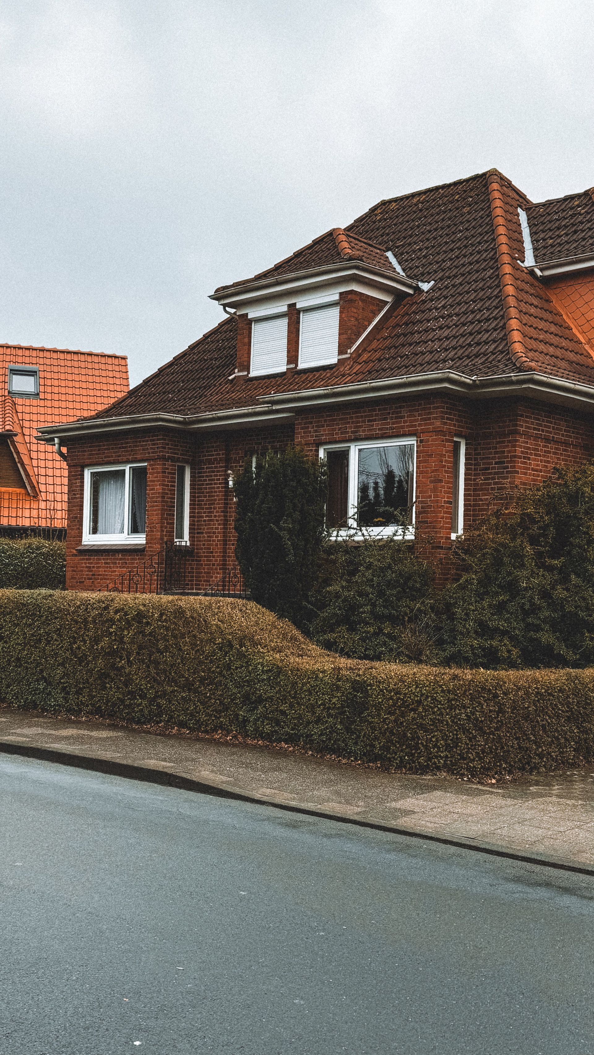 Brick house with a red-tiled roof beside a hedge along a residential street