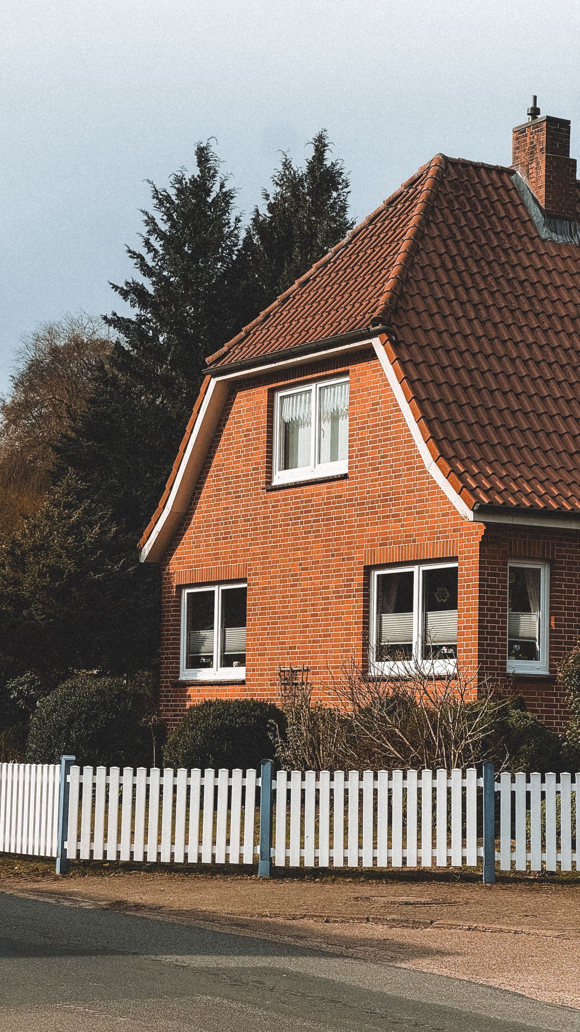 Red house with a red-tiled roof behind a white picket fence on a quiet street