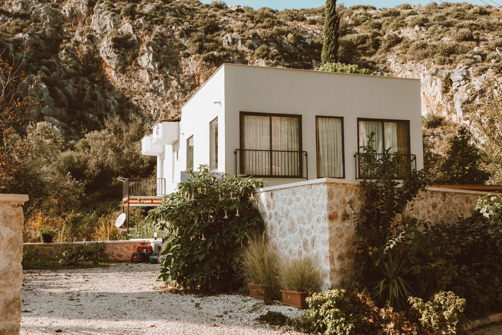 Small white house with a flat roof and surrounding plants against a rocky hillside