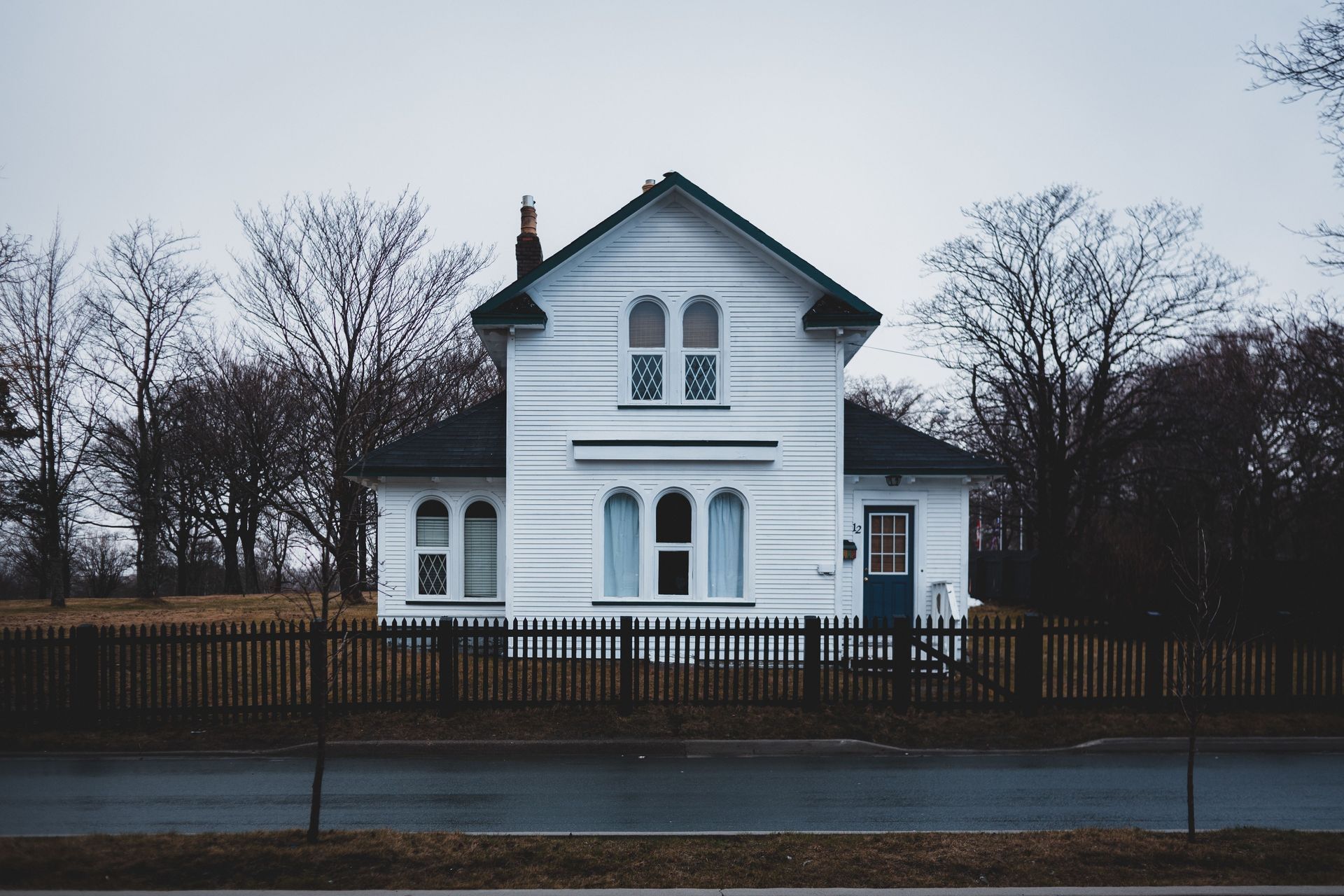White house with dark trim behind a fence on a rainy street, framed by leafless trees
