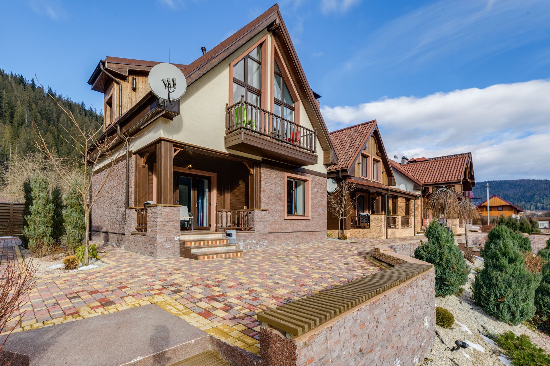 Modern mountain lodge with stone and wood exterior, red-tiled roofs, and a terrace overlooking hills and trees