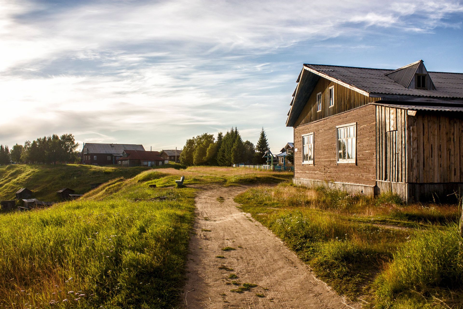 Rustic wooden house beside a dirt path in a sunny rural landscape with grassy hills and a cloudy sky