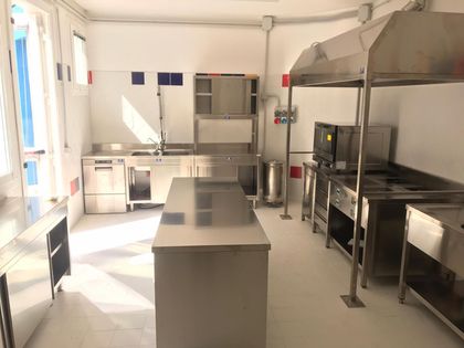 Cucina commerciale vuota con ripiani in acciaio inox, piano cottura, cappa e isola di preparazione in un ambiente luminoso.